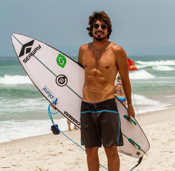 Gabriel Pastori na praia de óculos de sol e com uma prancha debaixo do braço
