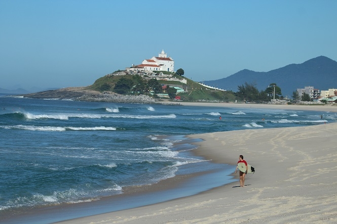 mulher caminhando na praia segurando uma prancha olhando pro mar