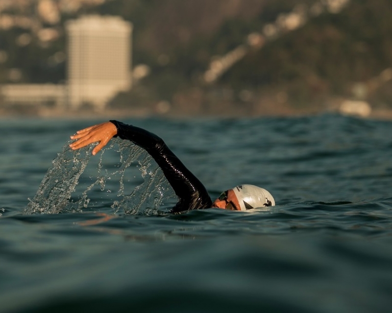 nadadora de touca branca e oculos de natacao no oceano com um predio desfocado ao fundo